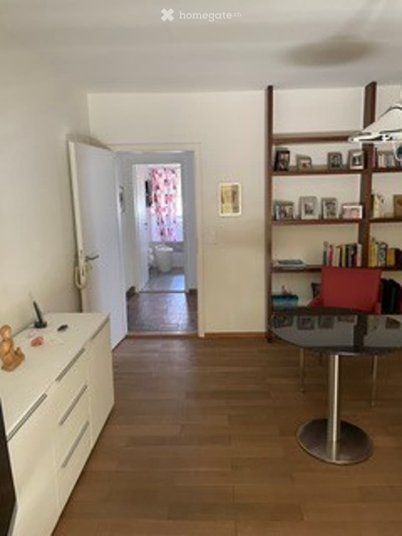 The image shows a modern, well-lit hallway leading to a room. The hallway features light wooden flooring, white walls, and a white cabinet with drawers on the left. A round glass table with a metal base and a red chair is positioned near a tall bookshelf