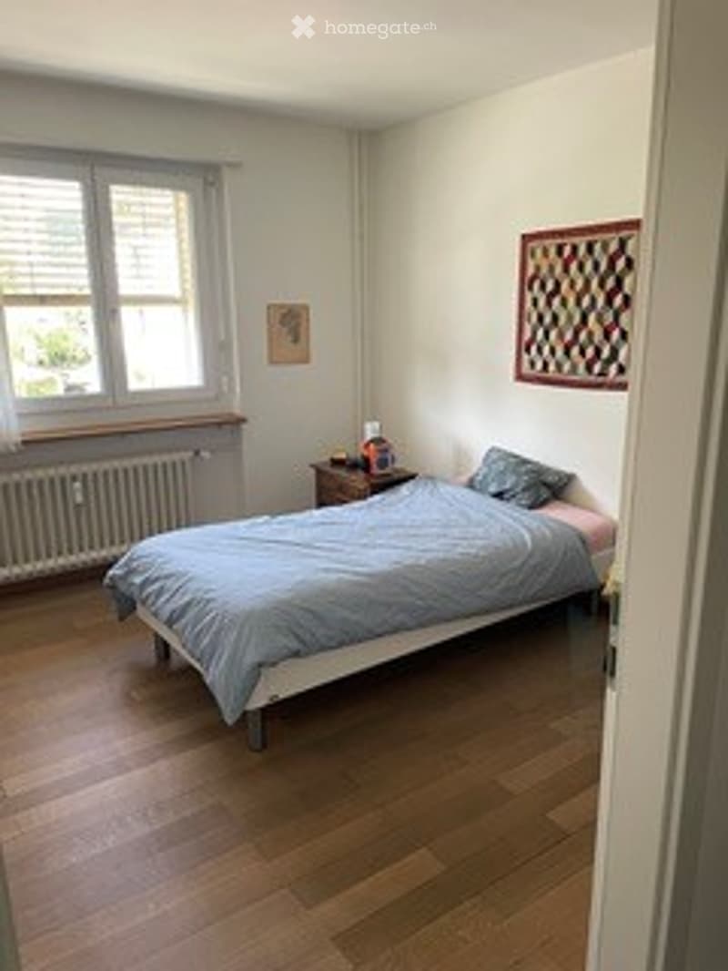 Cozy bedroom with wooden flooring, a single bed with blue bedding, a nightstand with a lamp, a radiator under the window, and a decorative picture frame on the wall.
