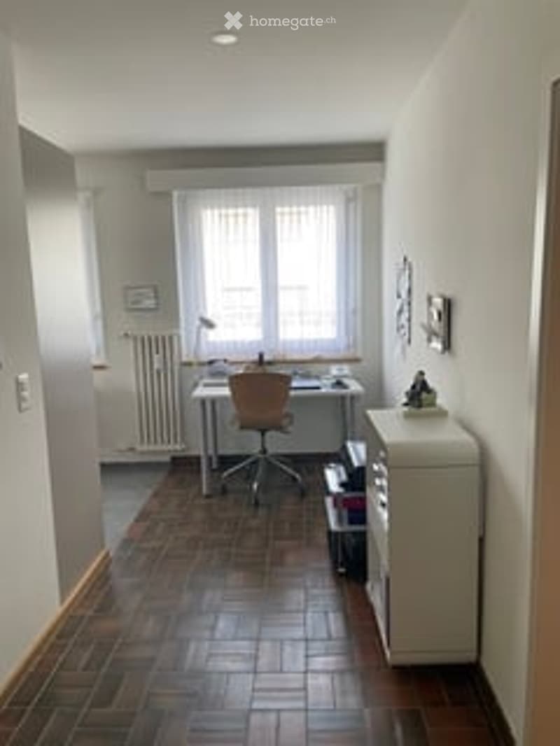 A small, well-lit room with wooden flooring, a large window, a desk with a chair, a lamp, a radiator, and a small cabinet.