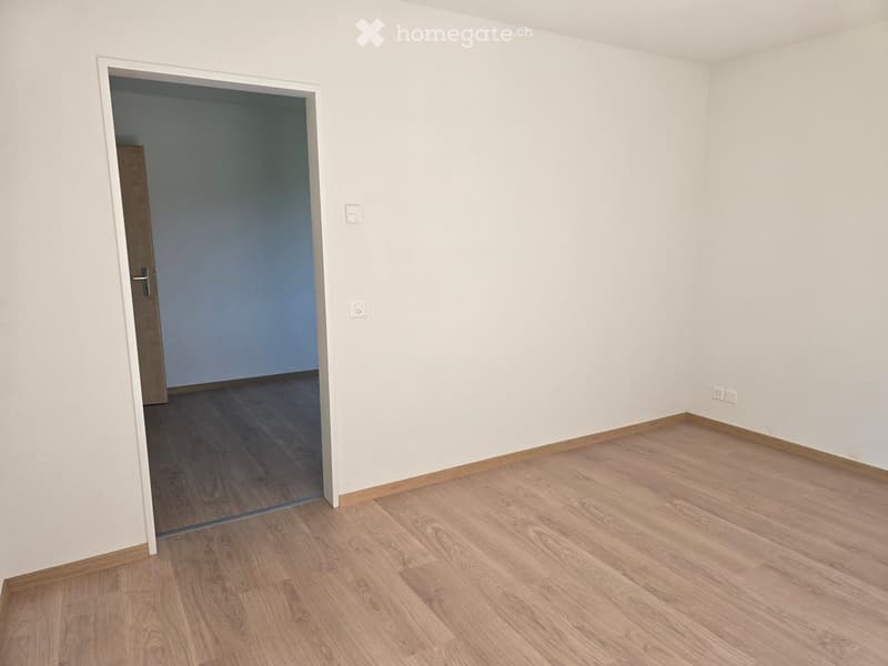 Empty room with light-colored walls and wooden flooring, featuring a doorway leading to another room.