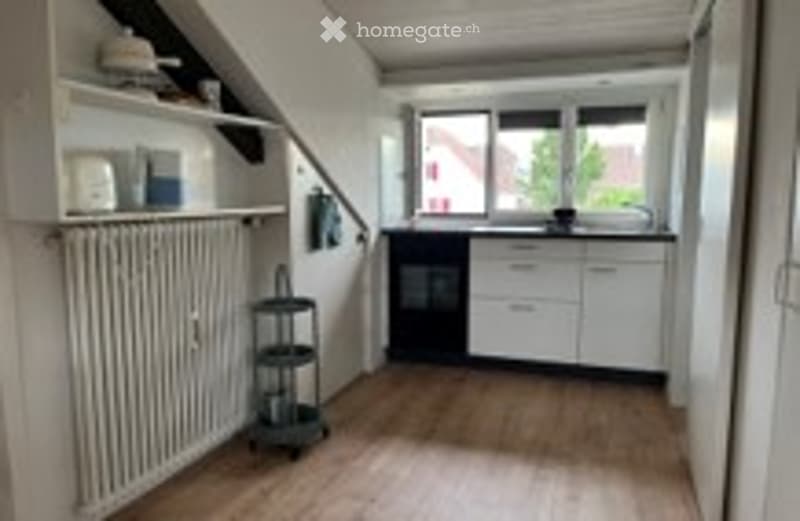 Bright, open-plan kitchen and living area with wooden flooring, white cabinets, a sink, radiator, and a small staircase leading to an upper level.