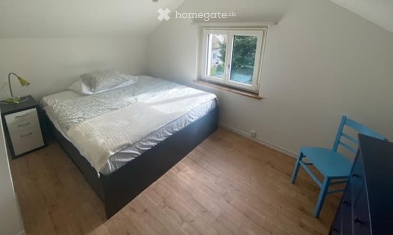 Cozy bedroom with wooden flooring, a bed with white bedding, a nightstand with a lamp, and a blue chair near the window.