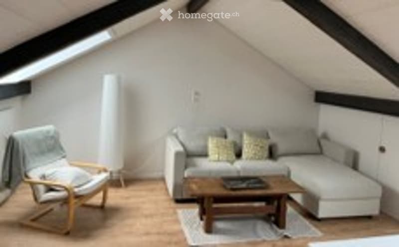 Cozy attic living space with exposed wooden beams, grey sectional sofa, wooden coffee table, white armchair, and floor lamp.