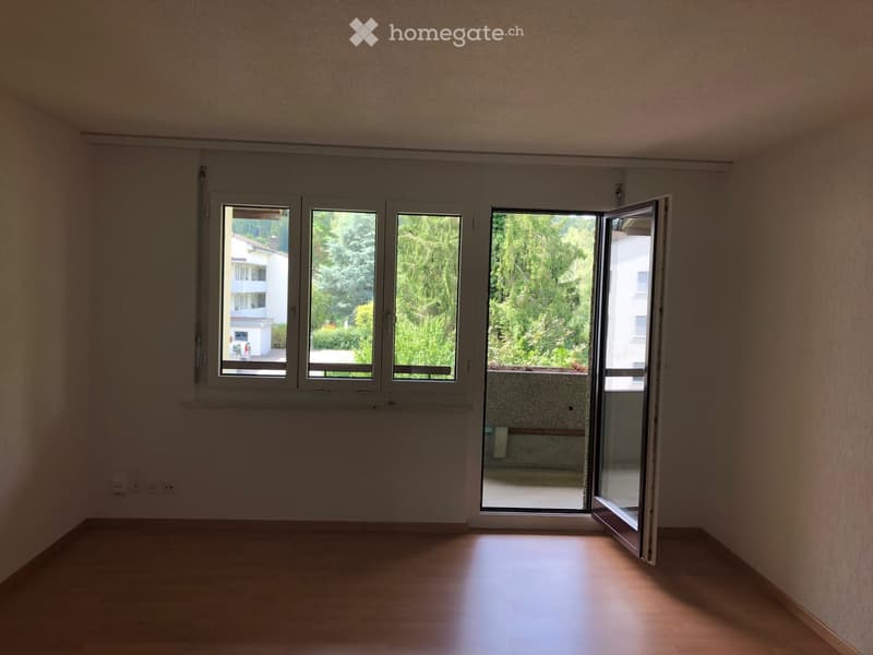 Empty room with wooden flooring, large windows, and an open sliding door leading to a balcony area with greenery visible outside.