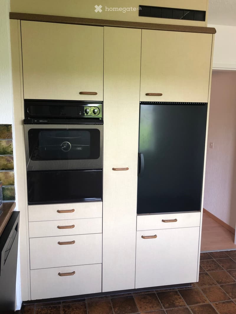 Modern kitchen with light-colored cabinets, stainless steel oven, and refrigerator. Wooden handles on cabinets, tiled floor.