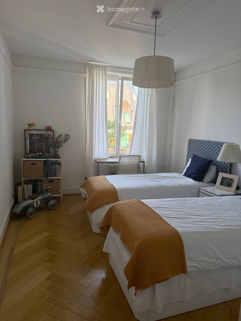 Bright bedroom with wooden flooring, two beds, nightstand, desk, chair, shelf, and decorative items.