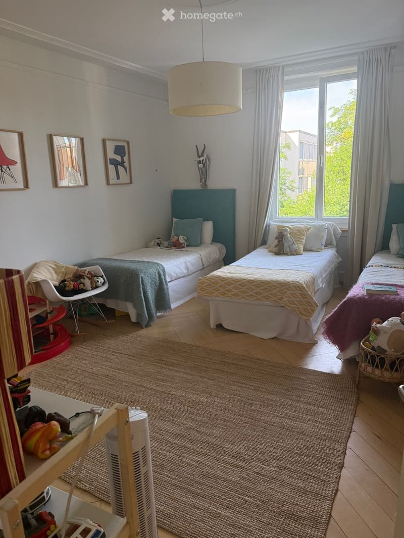Brightly lit children's bedroom with three single beds, each with colorful bedding and stuffed toys, a rocking chair, a small table with toys, and framed artwork on the walls.