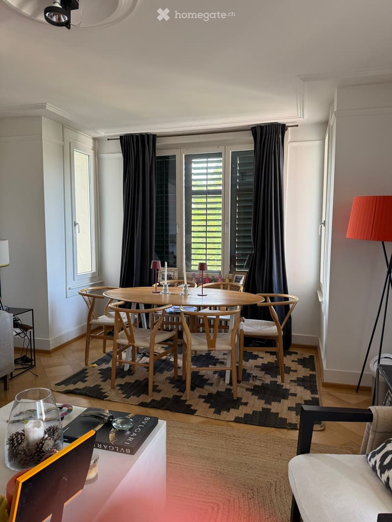 A modern, well-lit living and dining area with a round wooden dining table, six chairs, a sofa, a floor lamp, and decorative items on a side table.