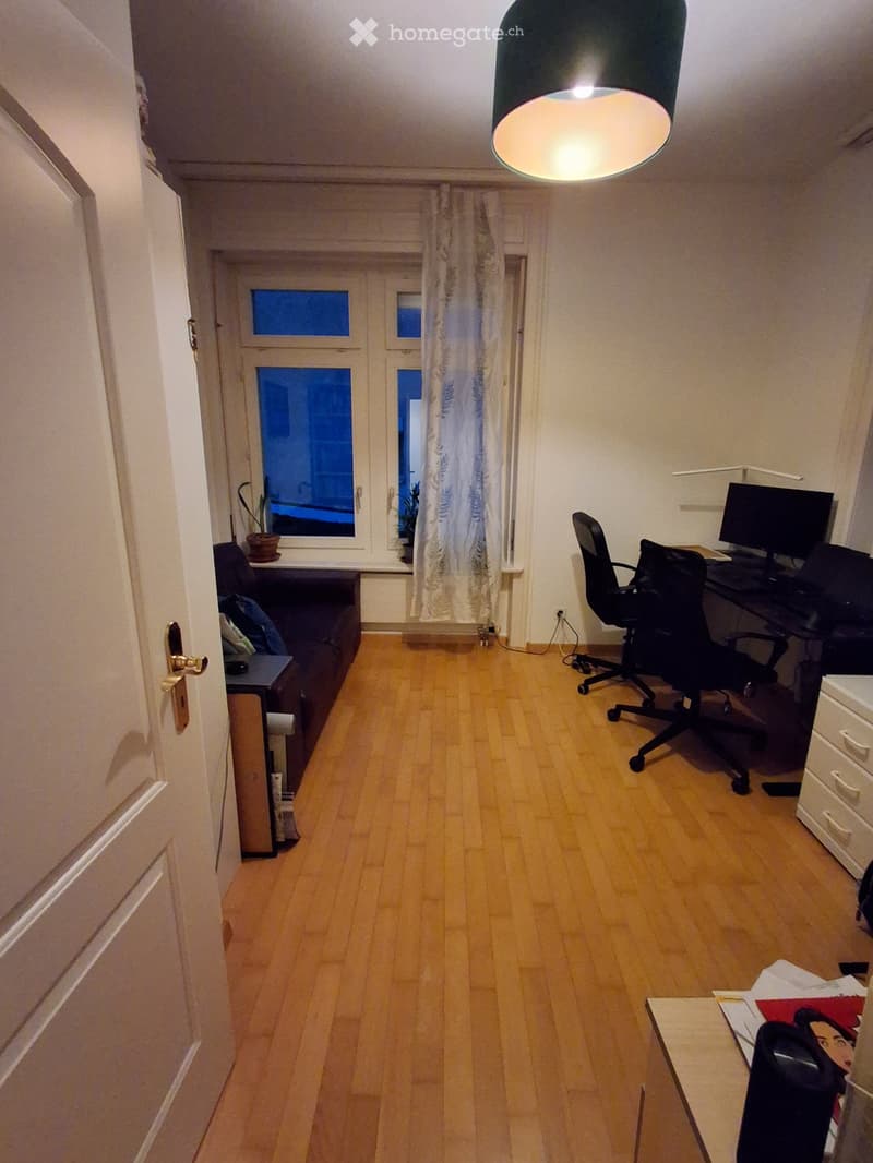 A furnished room with a wooden floor, a desk with a chair and screen, a sofa, a small table, and a window with curtains.
