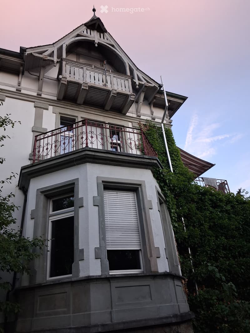 A multi-story house with white walls, ornate wooden balconies, and decorative railings. The building features large windows and a raised ground floor.