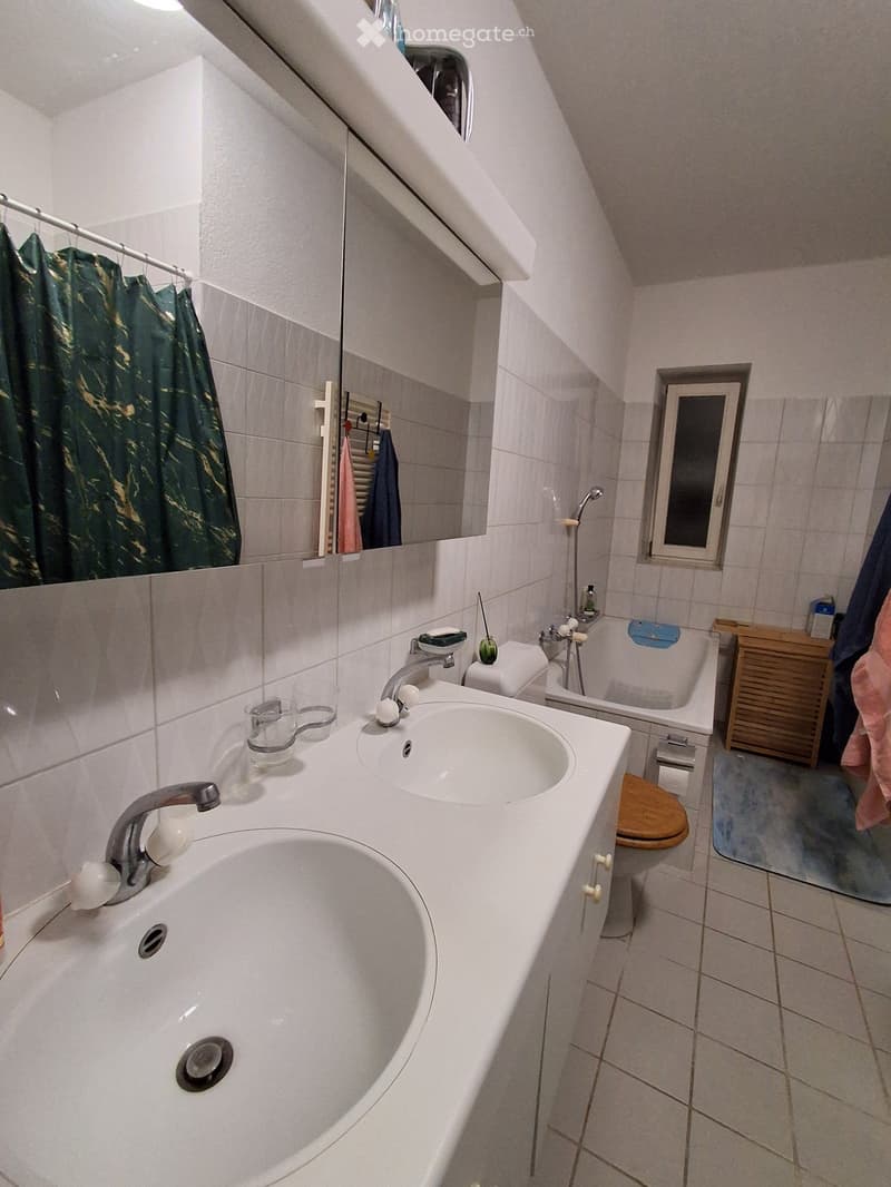 Bathroom with double sink, mirror, bathtub, toilet, and small window. Green shower curtain visible on the left.
