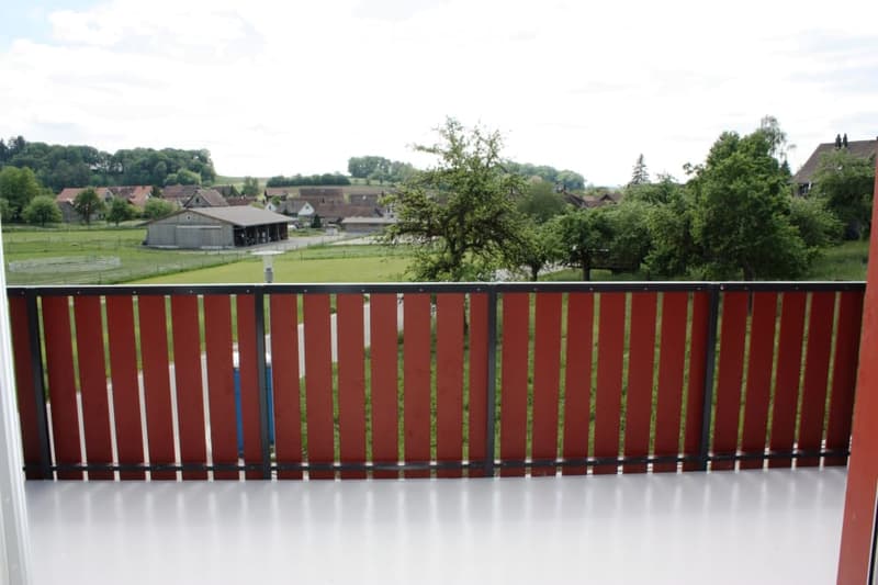 Balcony with red wooden railing, overlooking a rural landscape with green fields, trees, and distant buildings.