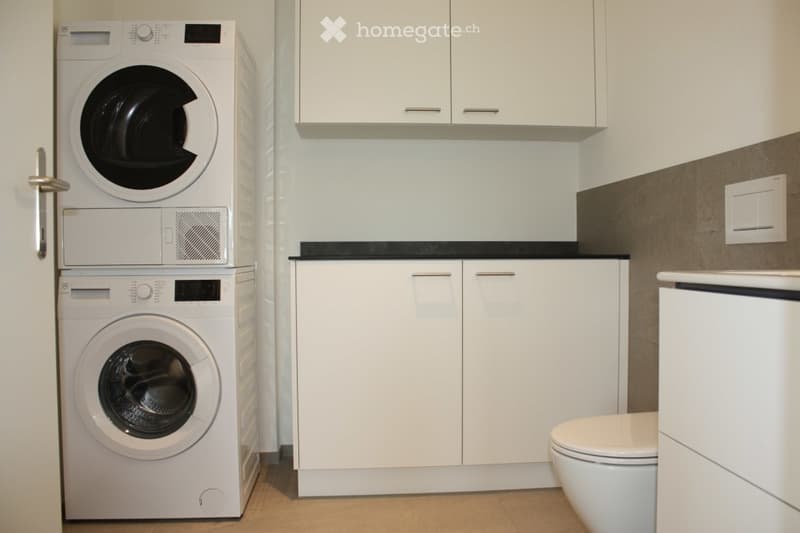 Modern bathroom with white cabinets, a black countertop, a toilet, and stacked washing machine and dryer.