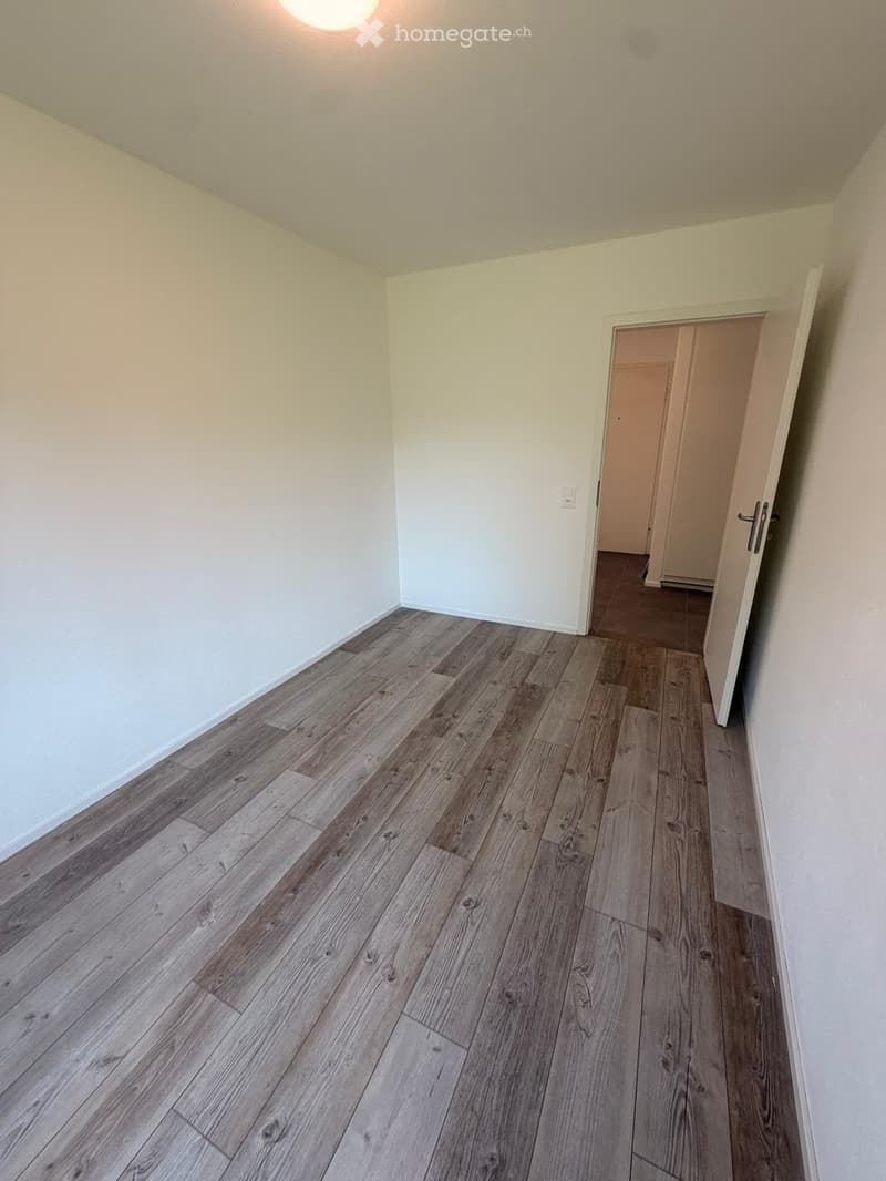 Empty hallway with wooden flooring and white walls, featuring an open door leading to another room.