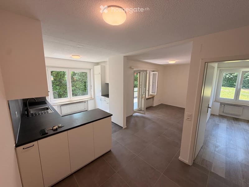 Modern kitchen with white cabinets, black countertop, and stainless steel sink. Large windows provide natural light, and the space is connected to a bright living area with tiled flooring.