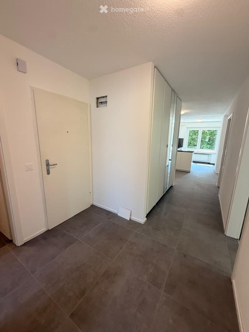 Empty hallway with tiled floor, white walls, and doors on both sides. The hallway leads to a room with windows and a visible kitchen area.