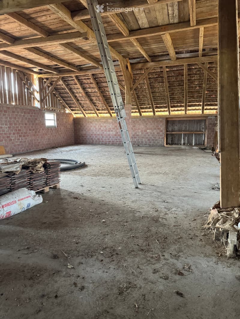 Empty attic space with exposed wooden beams, brick walls, and a concrete floor. Ladder and construction materials are present.
