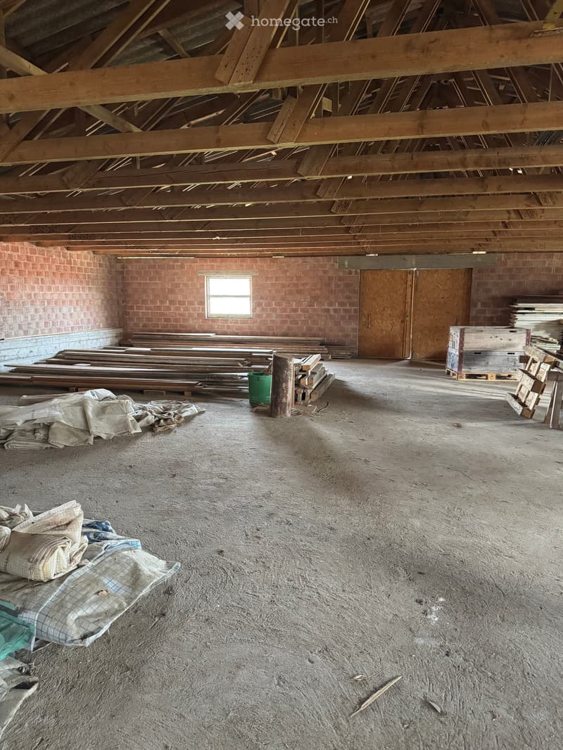 Empty attic space with exposed wooden beams, brick walls, a small window, and construction materials scattered around.
