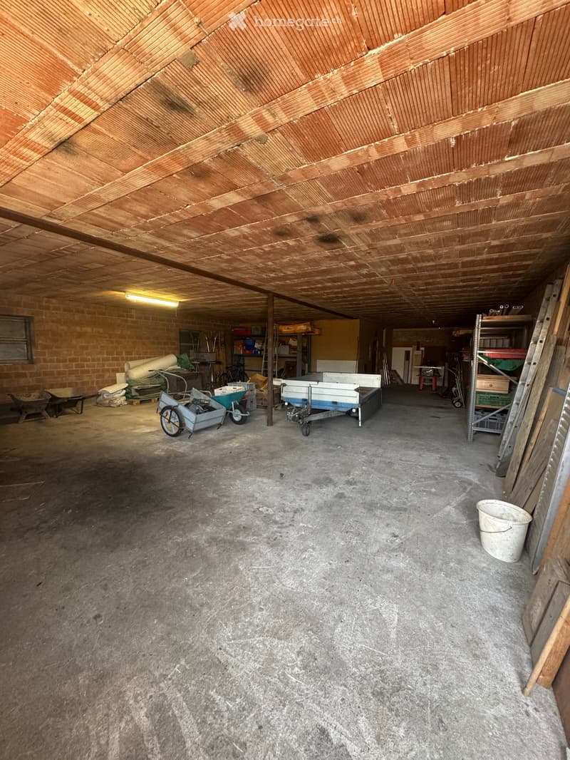 Empty garage with concrete floor, wooden ceiling, brick walls, and scattered tools and equipment.