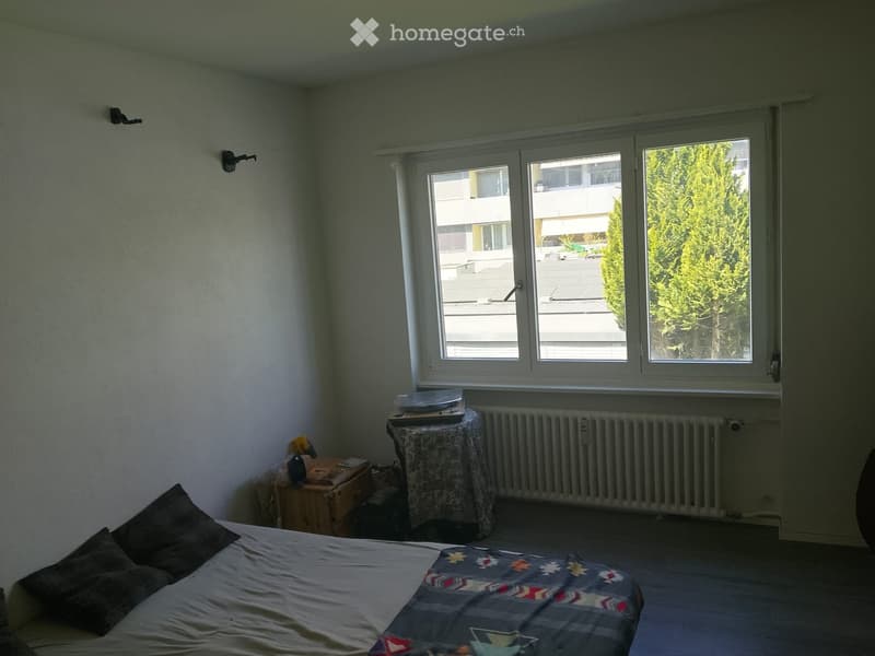 Bedroom with a bed, small table, radiator, and stool near the window.