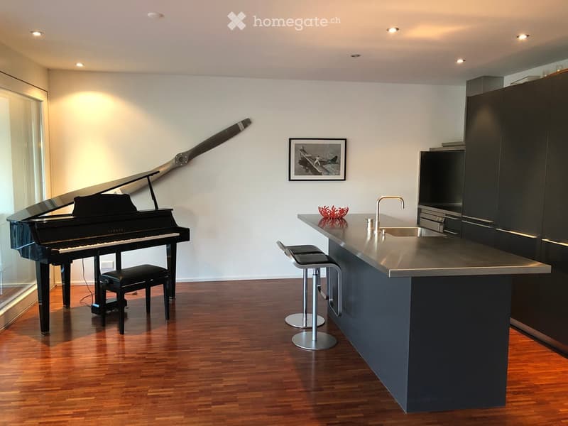 Modern room with a grand piano, black stool, kitchen island with sink and faucet, two bar stools, and a framed picture on the wall.