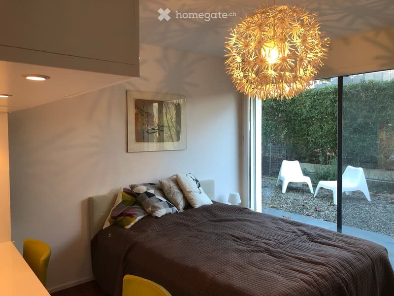 Modern bedroom with a large bed, colorful pillows, a yellow chair, a small table, a picture frame on the wall, and a decorative hanging light fixture above the bed. Large windows offer a view of the garden.