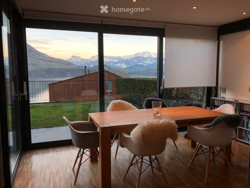 Dining area with a wooden table, chairs, and a large screen, overlooking a scenic mountain view through large glass doors.