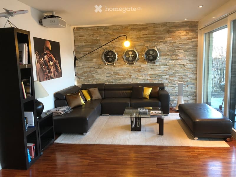 Modern living room with dark leather sectional sofa, glass coffee table, ottoman, bookshelf, wall-mounted lamps, and decorative clocks.