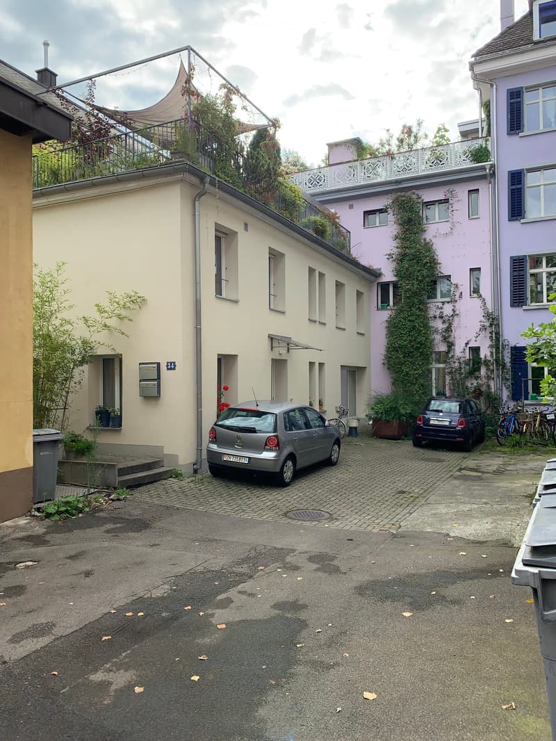 A small residential building with a balcony, surrounded by other buildings and parked cars in a narrow alleyway.