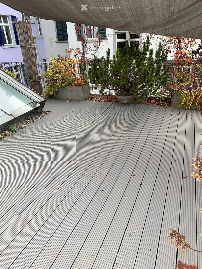 Wooden deck with potted plants and a canopy overhead, overlooking neighboring buildings.