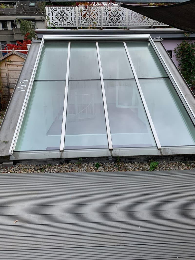 A modern glass skylight structure with a metal frame, situated on a flat, metal-roofed surface.