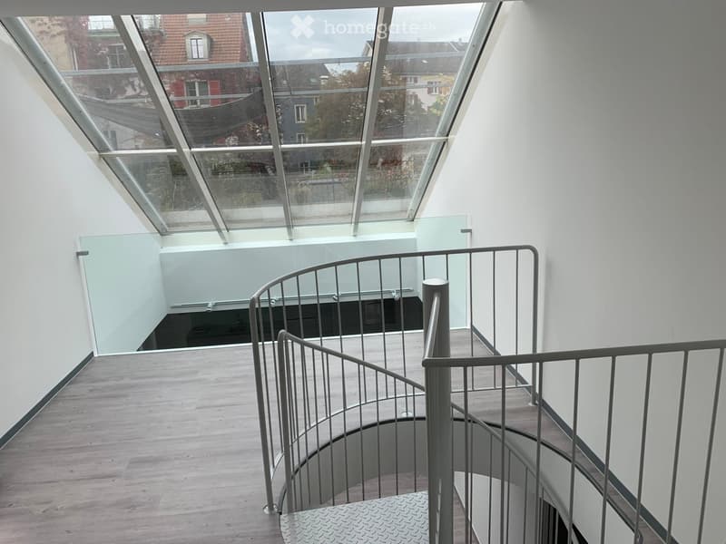 Spiral staircase with modern metal railing and glass ceiling, leading to an upper level with wooden flooring and white walls.