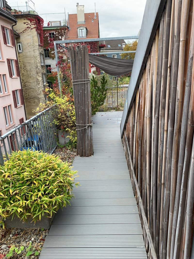 A narrow, elevated walkway with bamboo railings, surrounded by greenery and leading to a building entrance.