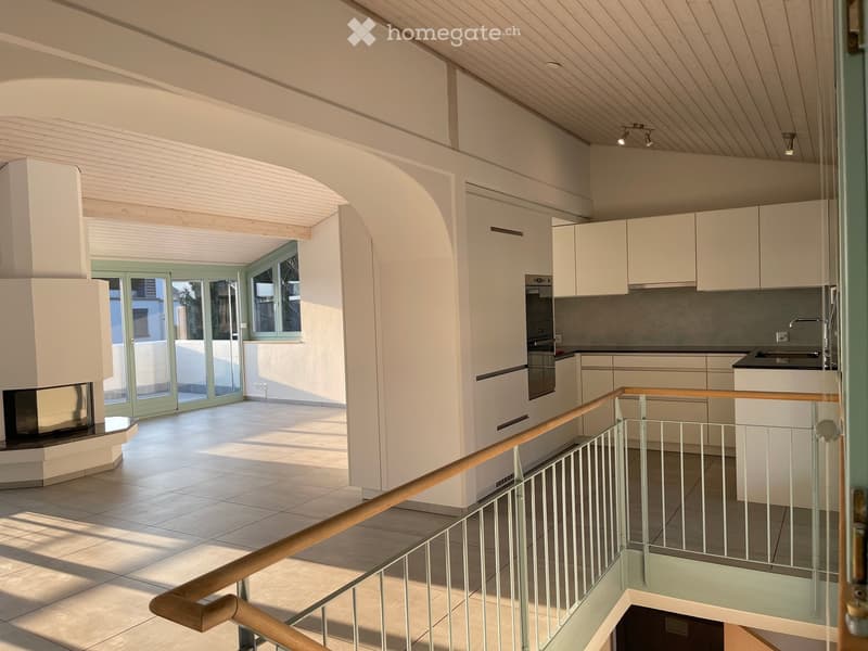 Modern, open-plan living and kitchen area with white cabinets, a central fireplace, and a staircase leading to an upper level.