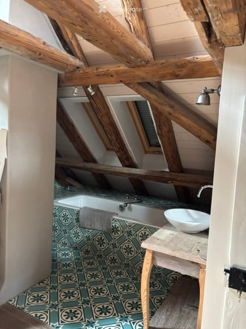 Bathroom with exposed wooden beams, green and white patterned tiles, a bathtub, a sink, and a small wooden stool.