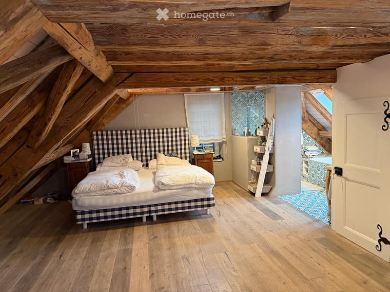Cozy attic bedroom with wooden beams, slanted ceilings, and wooden flooring. Features a double bed with a checkered headboard, nightstand, and a small shelf with decorative items.