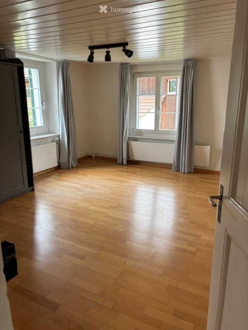 Empty room with wooden flooring, large windows with curtains, radiators, and track lighting.