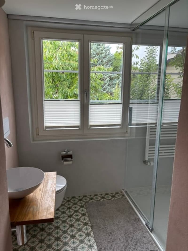 Modern bathroom with a wooden sink, toilet, and glass-enclosed shower. Features a window with blinds and a patterned tile floor.
