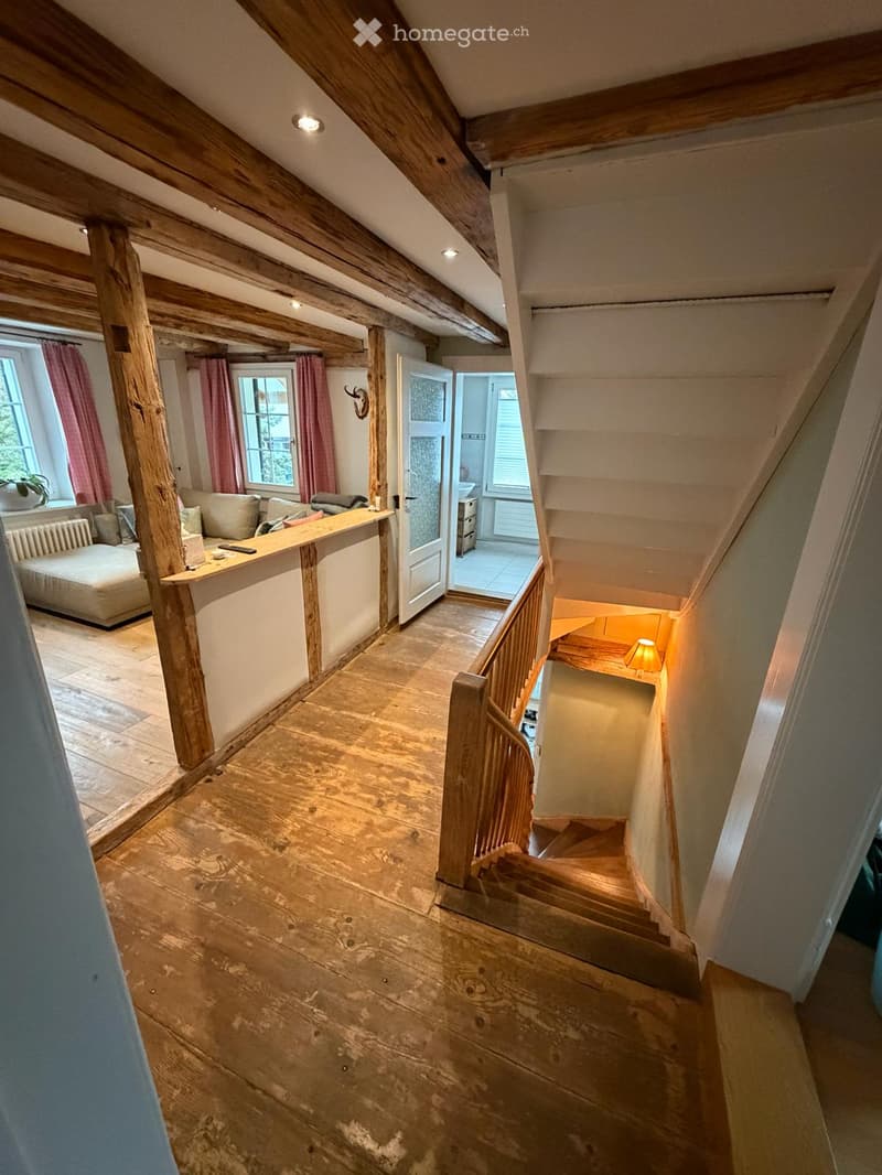 A rustic hallway with wooden beams, a staircase leading upwards, and a small seating area with a sofa and coffee table. The space features exposed wooden beams, wooden flooring, and large windows with red curtains.