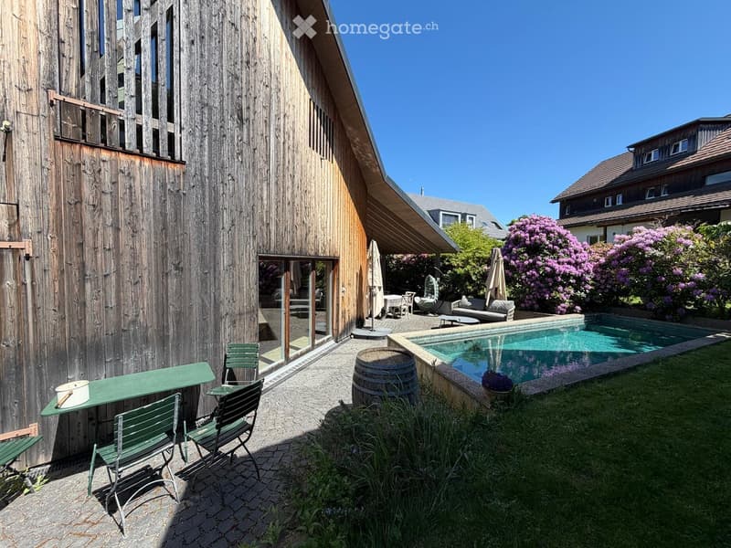 Outdoor garden area with a swimming pool, wooden decking, green metal chairs, a wooden barrel, and lush flowering bushes.