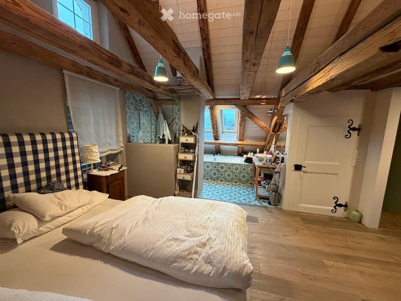 Cozy attic bedroom with wooden beams, exposed ceiling, large windows, and an en-suite bathroom featuring a bathtub and sink.