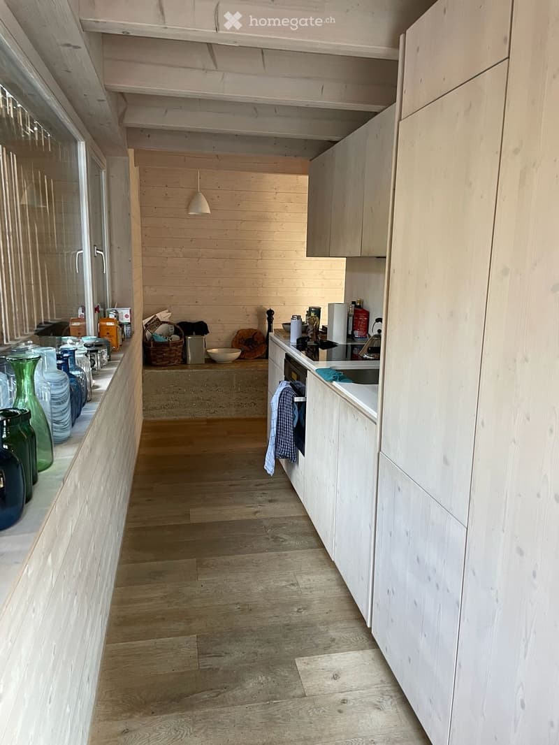 Modern kitchen with light wooden cabinets, a central island, and a variety of decorative items on the windowsill and counter. The space features a wooden floor and a hanging pendant light.
