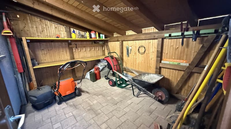 A well-organized garage with wooden walls and a slanted wooden ceiling. Contains a lawn mower, wheelbarrow, ladder, shelves with cleaning supplies, and various tools.