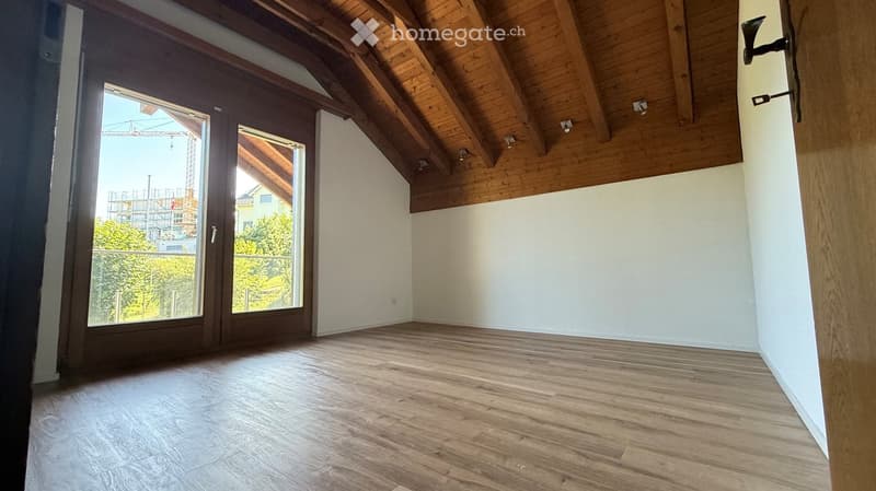 Empty room with wooden flooring, white walls, and exposed wooden beams on the ceiling. Large sliding glass doors lead to a balcony with a view of greenery and a construction site.