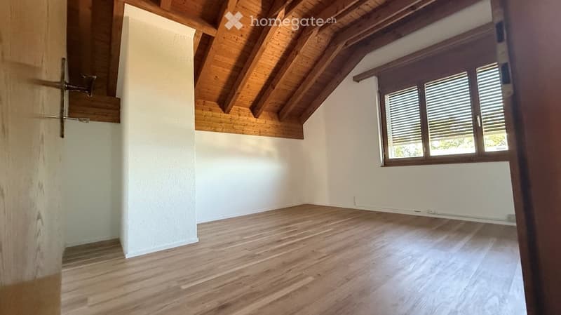 Empty room with wooden flooring, white walls, and exposed wooden ceiling beams. Features a window with blinds and a small staircase leading upwards.