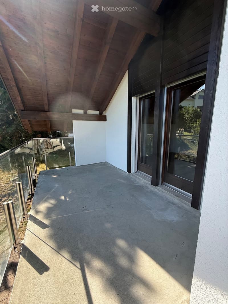 Modern balcony with a wooden roof, glass railing, and concrete floor, featuring large windows and a view of the surrounding greenery.