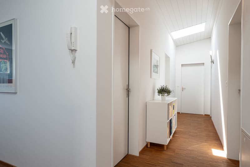 Modern hallway with light-colored walls, wooden flooring, and built-in shelving unit with books and a small plant. Features a wall-mounted telephone and framed artwork.