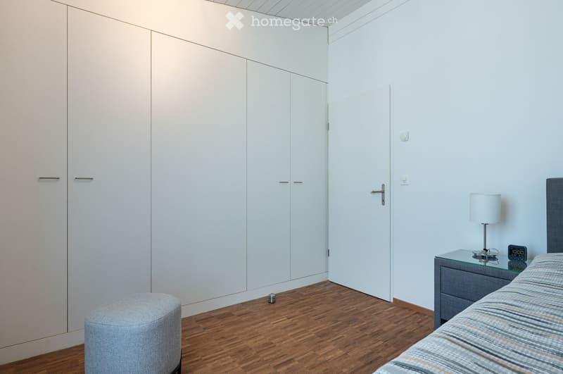Modern bedroom with a grey bed, nightstand, lamp, and a grey ottoman. The room features light-colored walls, wooden flooring, and built-in white wardrobes.