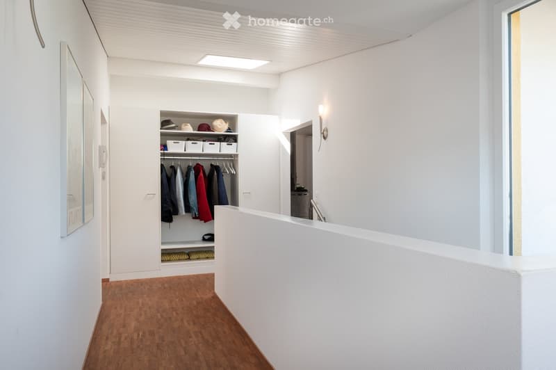 Modern, minimalist hallway with white walls, wooden flooring, built-in closet with clothing and storage boxes, and a sleek white reception desk.