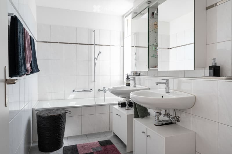 Modern bathroom with white tiles, a bathtub, two sinks, a large mirror, and a wicker basket on the floor.